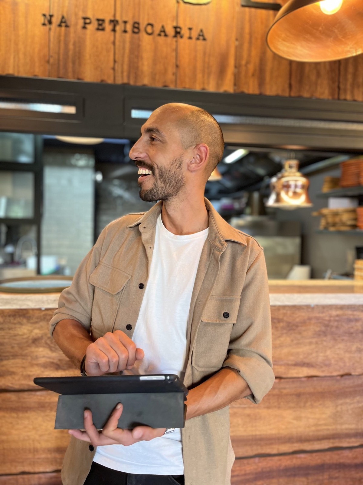 Host do Banzé em frente à cozinha aberta, com o letreiro «Na Petiscaria» gravado na madeira